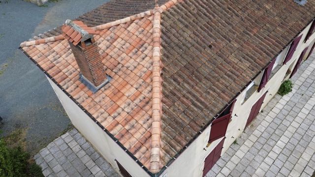 Vue aérienne d'une toiture en tuiles mécaniques en cours de nettoyage : la partie gauche est nettoyée, la partie droite est encore couverte de dépôts sombres
